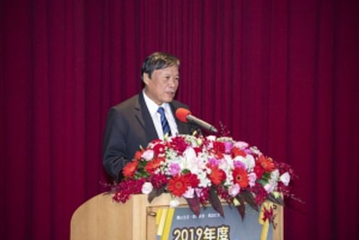 A gentleman at speaker's desk in front of a red curtain and red and white flowers