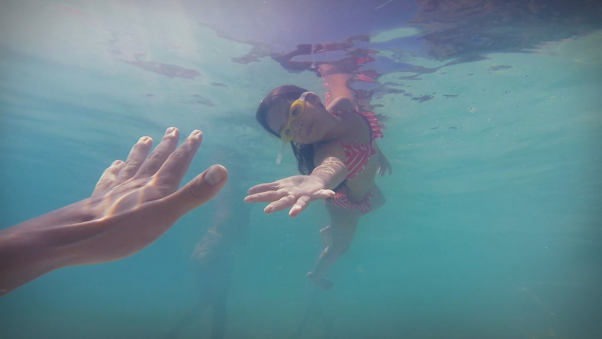 Girl diving in water with diving goggles and grabbing outstretched hand under water.